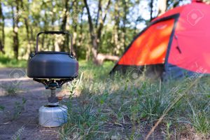 Close-up image burning gas stove and kettle boiling water located green grass lawn outdoor red tent focus on stove and fire tent is blurred